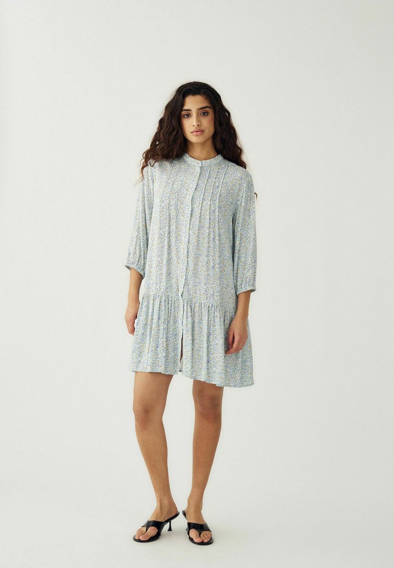 Kobieta o długich kręconych włosach, ubrana w jasnoniebieską sukienkę w kwiaty z plisowaniem i czarne sandały na obcasie, stoi na tle jednolitego białego tła.