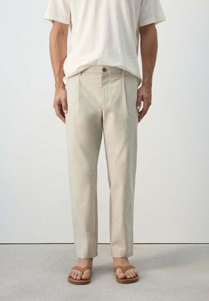 Mann trägt beige gerade geschnittene Hose, weißes Kurzarmhemd und braune Sandalen und steht auf einem grauen Boden vor einer schlichten Wand.