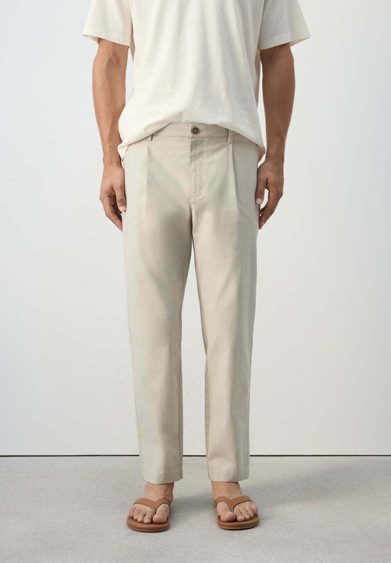 Homme portant un pantalon beige coupe droite, une chemise blanche à manches courtes et des sandales marron, debout sur un sol gris devant un mur uni.