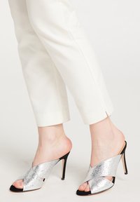 Mule metalice argintii cu vârful deschis, cu un design încrucișat, talpi negre și toc subțire, asortate cu pantaloni albi scurtați.