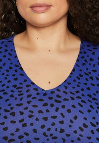 Kék anyagból készült V-nyakú póló fekete leopárd foltokkal. Simán tapintású, testhezálló dizájn, amely kiemeli a nyakat és a vállakat.