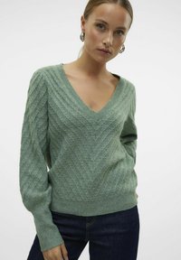 Maglione verde lavorato a maglia con scollatura a V, motivo testurizzato e maniche lunghe, abbinato a jeans scuri. Presenta polsini e orlo a coste.