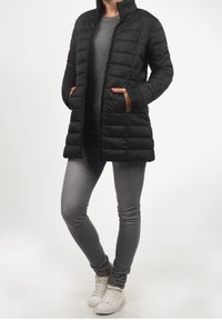 Černý prošívaný kabát na zip s bočními kapsami a vysokým límcem, doplněný šedými úzkými džínami a bílými teniskami. Hladká textura látky.