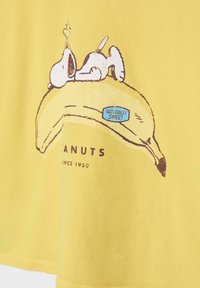 Snoopy ligger på en stor gul banan med en "Naturligt Sød" klistermærke, en del af et Peanuts-tema grafik på gul stof.