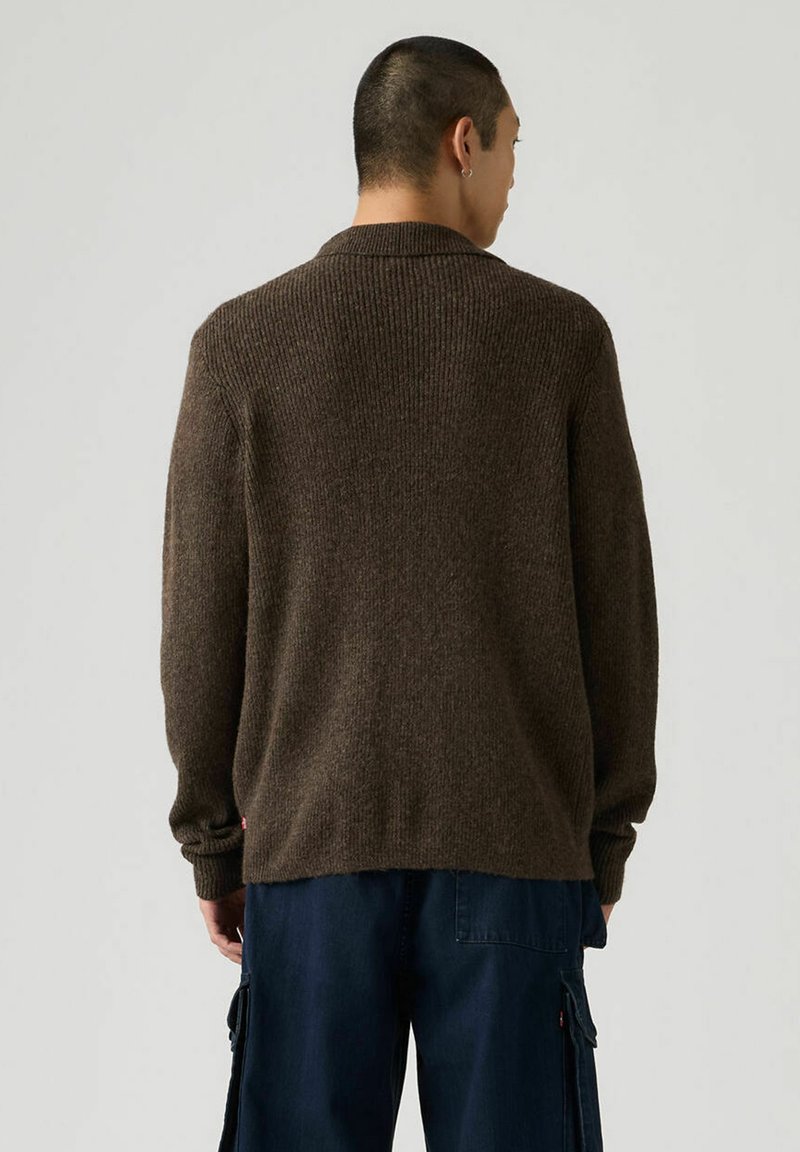 Pull oversize en maille côtelée marron avec un col châle, des manches longues et une coupe décontractée, associé à un pantalon cargo bleu foncé.