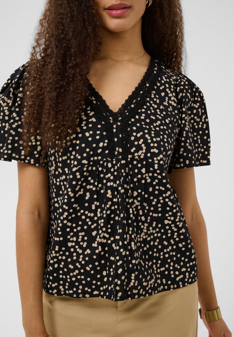 Femme aux cheveux bouclés portant un chemisier noir col V à pois roses et beiges et un pantalon beige sur un fond uni.