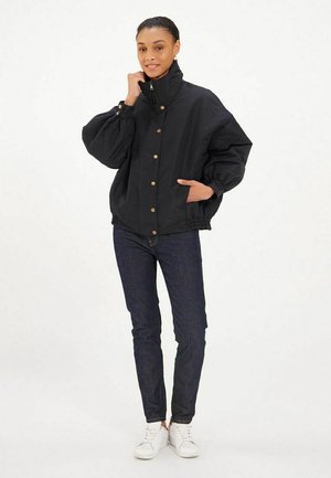 Veste noire surdimensionnée avec un col haut, des boutons et des poignets élastiques, associée à un jean foncé et des baskets blanches.