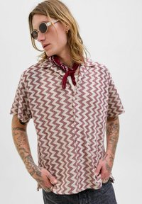 Camicia a maniche corte con bottoni, a motivo zigzag in bordeaux e crema, realizzata in tessuto leggero, indossata con un bandana rossa.
