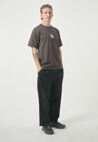 Brown cotton t-shirt with a purple graphic design on the chest, paired with loose-fitting black pants and black sneakers.