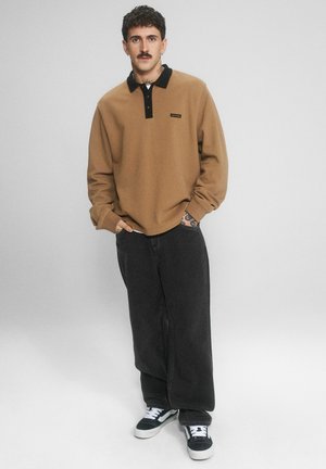 Mężczyzna z wąsami i tatuażami, ubrany w brązowe polo z długim rękawem, szerokie czarne spodnie oraz czarno-białe tenisówki, stojący z rękami w kieszeniach.