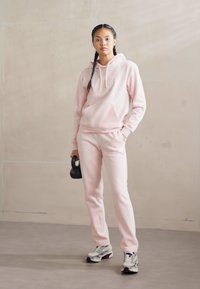 Camisola cor-de-rosa com um bolso canguru, combinada com calças de jogging a condizer. O modelo segura um kettlebell preto, vestindo ténis elegantes.