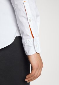 Biała koszula z długim rękawem i mankietem na guziki, z pomarańczowym akcentem wzdłuż boku. Gładka faktura, klasyczny kołnierzyk.