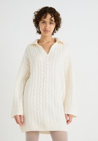 Valge kootud sweater'i kleit, millel on kabriku muster, kaelusega v-kaelus ja pikad varrukad. Pehme, tekstureeritud kangas.