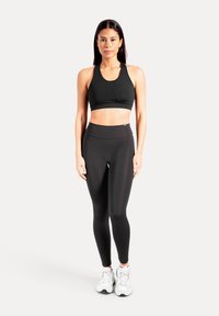 Reggiseno sportivo nero e leggings a vita alta in tessuto liscio. Modella indossa scarpe da ginnastica bianche. Silhouette semplice e atletica con un design aderente.