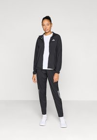 Ensemble veste et pantalon de sport noirs en matériau léger avec bandes réfléchissantes sur le pantalon, assorti à des chaussures de sport blanches.
