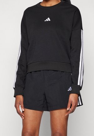 Persoon draagt een zwarte longsleeve top met witte strepen op de mouwen en zwarte korte broek met een logo op de linker dij.