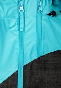Giacca turchese con tessuto scuro a contrasto, zip verticali e una superficie liscia caratterizzata da cuciture angolate e rinforzate.