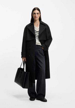 Donna che indossa un lungo cappotto nero, maglione bianco e nero a motivo, pantaloni neri larghi, con una grande borsa nera in mano, su uno sfondo semplice.