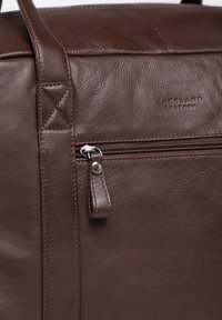 Brown leather bag featuring a zippered pocket with a leather pull tab, detailed stitching, and embossed brand logo. Smooth texture and structured design.