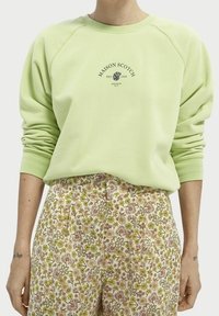 Persoon draagt een lichtgroene Maison Scotch-trui en een hooggetailleerde bloemenbroek in tinten roze, groen en bruin.