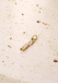 Goldschleifenanhänger mit zwei unregelmäßig geformten, schimmernden weißen Perlen und einem kleinen goldenen Perlenakzent auf einer strukturierten hellen Oberfläche.