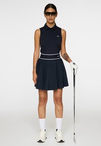 Nainen, joka käyttää ilman hihoja tummansinistä golf-asua, valkoista hanskaa, aurinkolaseja ja pitää golf mailaa kädessään seisten yksiväristä taustaa vasten.