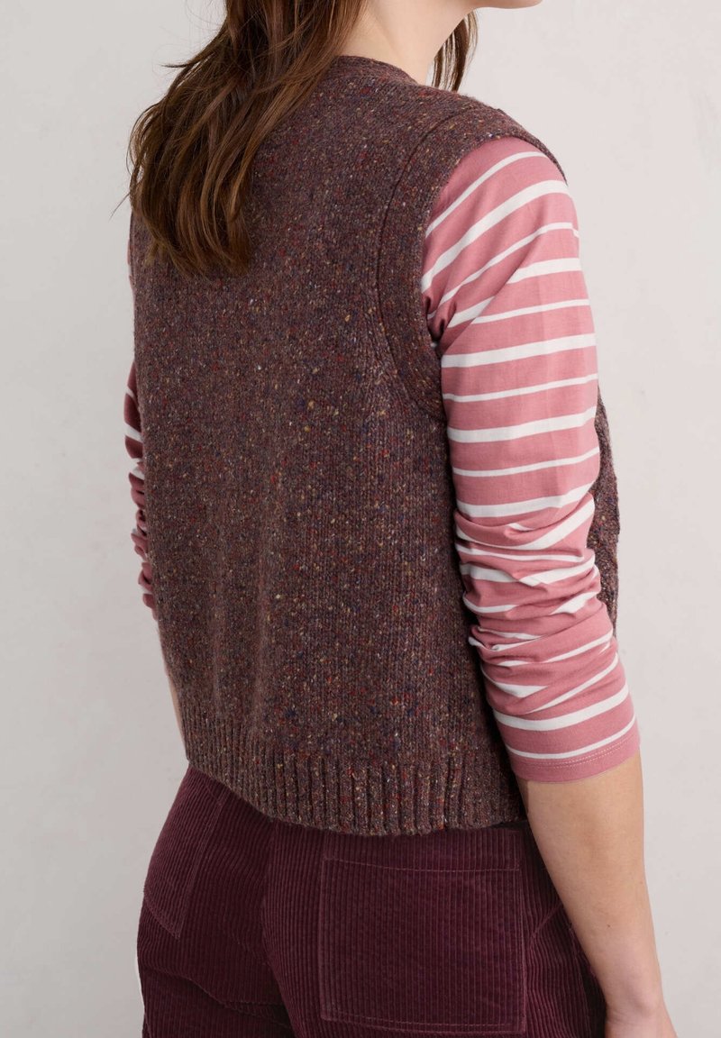 Kobieta ubrana w brązową, nakrapianą dzianinową kamizelkę na różowo-biale paskowanej koszuli z długim rękawem oraz bordowe spodnie sztruksowe, widziana od tyłu.