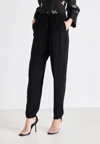 Pantalons noirs amples avec une taille à cordon de serrage, des poches latérales et des jambes fuselées, associés à des talons hauts noirs à lanières.