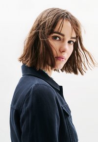 Tummansinisestä denimpuvusta valmistettu takki, jossa on kauluksen malli, rintatasku ja näkyvä ompelu. Mallilla on lyhyet ruskeat hiukset ja neutraali ilme.