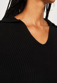 Gros plan d'une personne portant un pull noir texturé à col en V, avec de longues tresses et une grande boucle d'oreille en or.