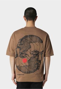 Brun kortærmet t-shirt med et line art design af et ansigt og en rød cirkel på ryggen. Blødt stof med en afslappet pasform.