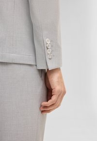 Main d'homme reposant à côté de la manche d'une veste de costume gris clair avec quatre boutons blancs dans un cadre intérieur neutre.