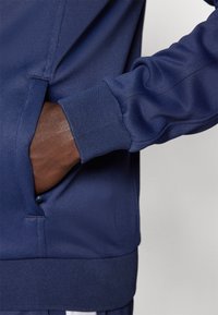 Hand in der Tasche einer dunkelblauen Jacke mit geripptem Bündchen und Saum, Nahaufnahme der Ärmel- und Seitennahtdetails.