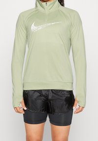 Ljusgrön långärmad träningströja med halv dragkedja och vit Nike-logotyp; kombinerad med svarta shorts som har en strömlinjeformad design och tygtextur.