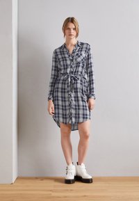 Abito camicia a quadri blu navy e bianco con maniche lunghe, chiusura con bottoni, vita svasata e orlo curvo. Indossato con stivali bianchi con tacco spesso.