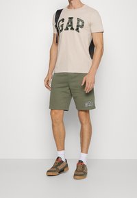 Camiseta de algodón beige con logo "GAP" en camuflaje verde, combinada con pantalones cortos verde oliva con un detalle blanco "GAP". Zapatos marrones con detalles en negro y rojo.
