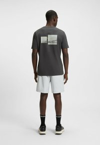 Camiseta gris de manga corta con dos gráficos rectangulares en la parte de atrás. Combinada con pantalones cortos claros y zapatillas negras.