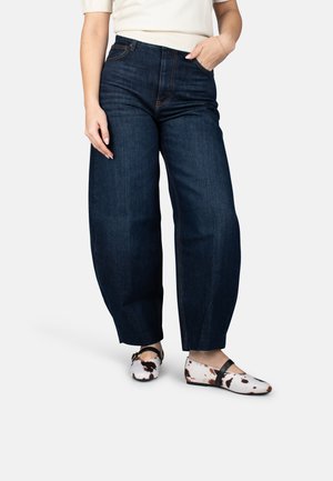 Mujer vestida con jeans oscuros de pierna ancha, camiseta blanca, una mano en el bolsillo y zapatos Mary Jane con estampado de vaca y tiras negras.