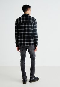 Chemise en flanelle à carreaux noir et blanc, à manches longues, coupe décontractée, associée à un jean gris et des chaussures noires, vue de dos.
