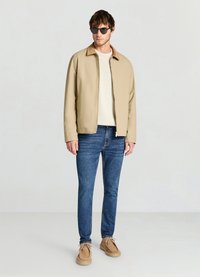 Veste légère beige, pull crème, jean bleu et chaussures marron clair. Les lunettes de soleil ajoutent une touche moderne ; design simple mais fonctionnel avec une texture minimale.