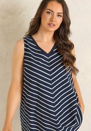 Woman with long wavy brown hair wearing a sleeveless navy blue dress with white diagonal stripes, standing against a beige background.