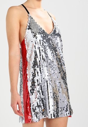 Kobieta w bezrękawnym srebrnym miniaturowym sukience z cekinami, z cienkimi czarnymi ramiączkami i czerwonymi cekinowymi panelami po bokach, na jednolitym tle.