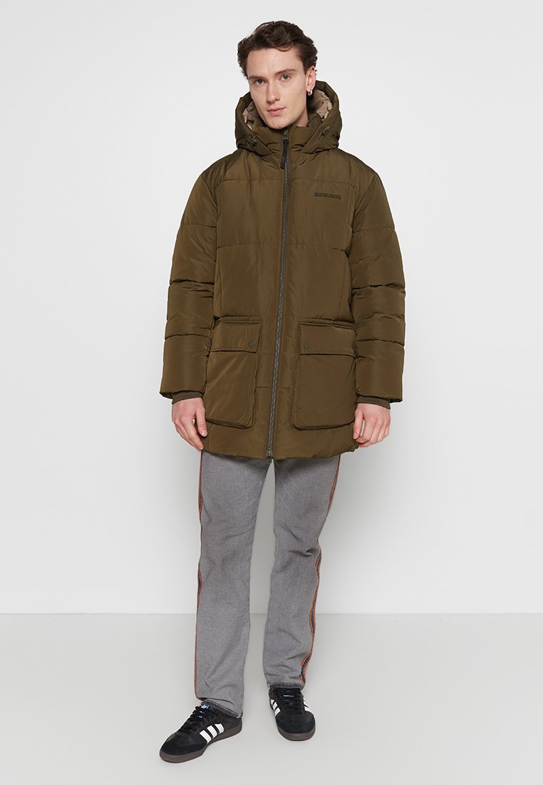 Mann trägt eine olivgrüne, kapuzenverstellbare Daunenjacke, graue Hose mit orangenen Streifen und schwarze Sneakers, während er vor einem schlichten weißen Hintergrund steht.