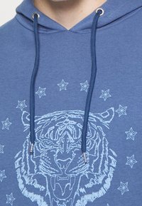 Blaues Kapuzenpullover mit einem Tiger-Gesichtsdesign in Hellblau, das mit Sternakzenten um das Motiv herum versehen ist und Kordeln mit metallenen Enden hat.