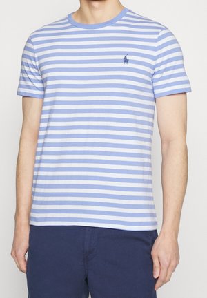 Mężczyzna noszący jasnoniebieską i białą koszulkę w poziome paski na krótki rękaw z małym haftowanym logo na piersi, w połączeniu z granatowymi spodniami.