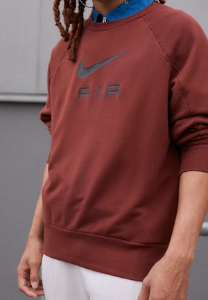 Person wearing rust-colored Nike Air sweatshirt with black logo, layered over blue collared shirt and paired with white pants.