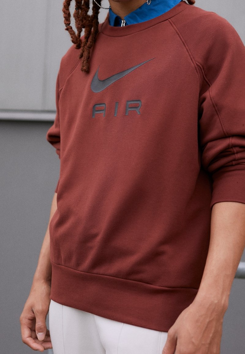 Personne portant un sweat-shirt Nike Air couleur rouille avec un logo noir, superposé à une chemise bleue à col et associé à un pantalon blanc.
