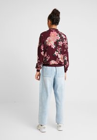 Kwiatowa kurtka bomber w ciemnym burgundzie z różowymi i zielonymi wzorami, zestawiona z jasnoniebieskimi spodniami o wysokim stanie. Posiada elastyczne mankiety i pas.