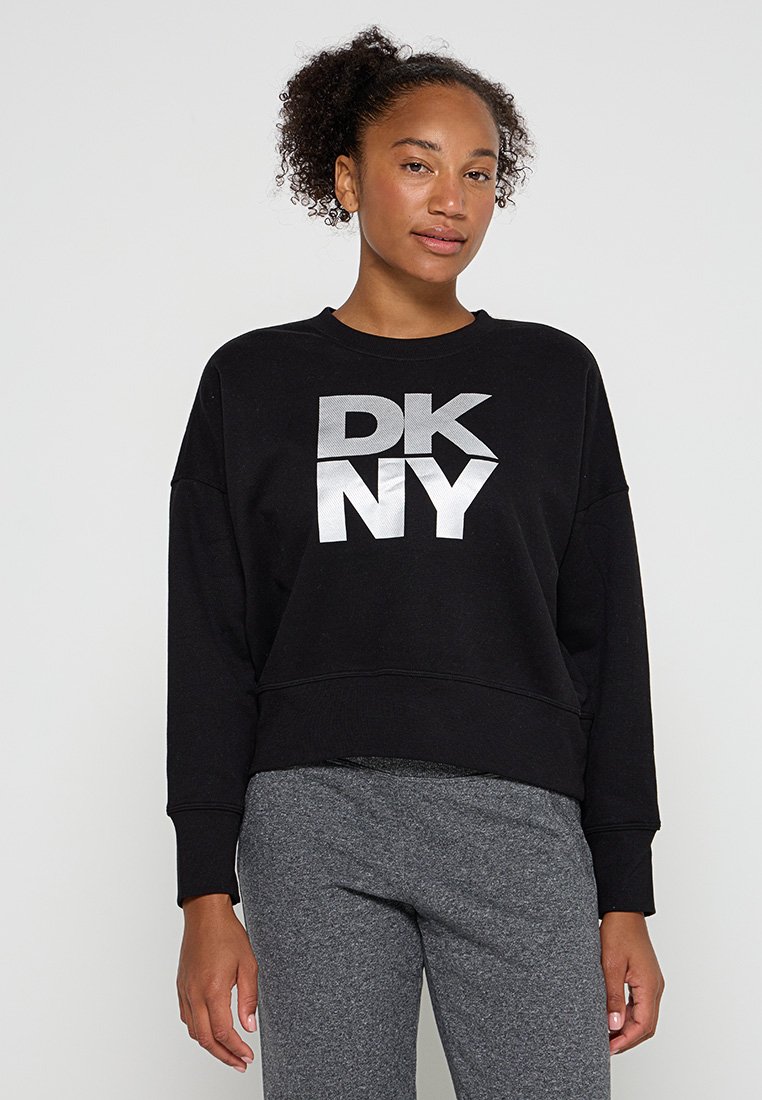 Junge Frau mit lockigem Haar, die einen schwarzen DKNY-Pullover und graue Hosen trägt, stehend vor einem schlichten weißen Hintergrund.