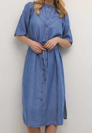 Femme portant une robe en denim bleue mi-longue avec des boutons sur le devant et une ceinture nouée à la taille, debout devant un fond clair uni.